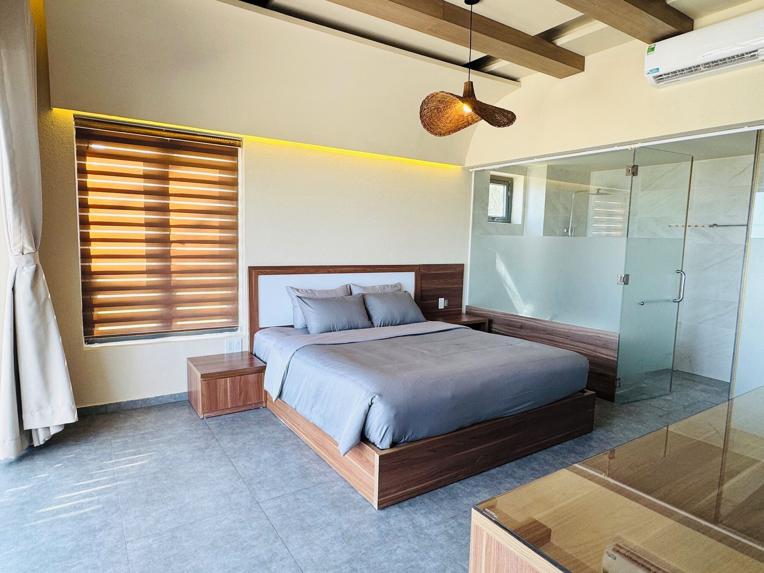 A modern bedroom with a wooden bed, a bedside table, a glass wardrobe, and a window with blinds.