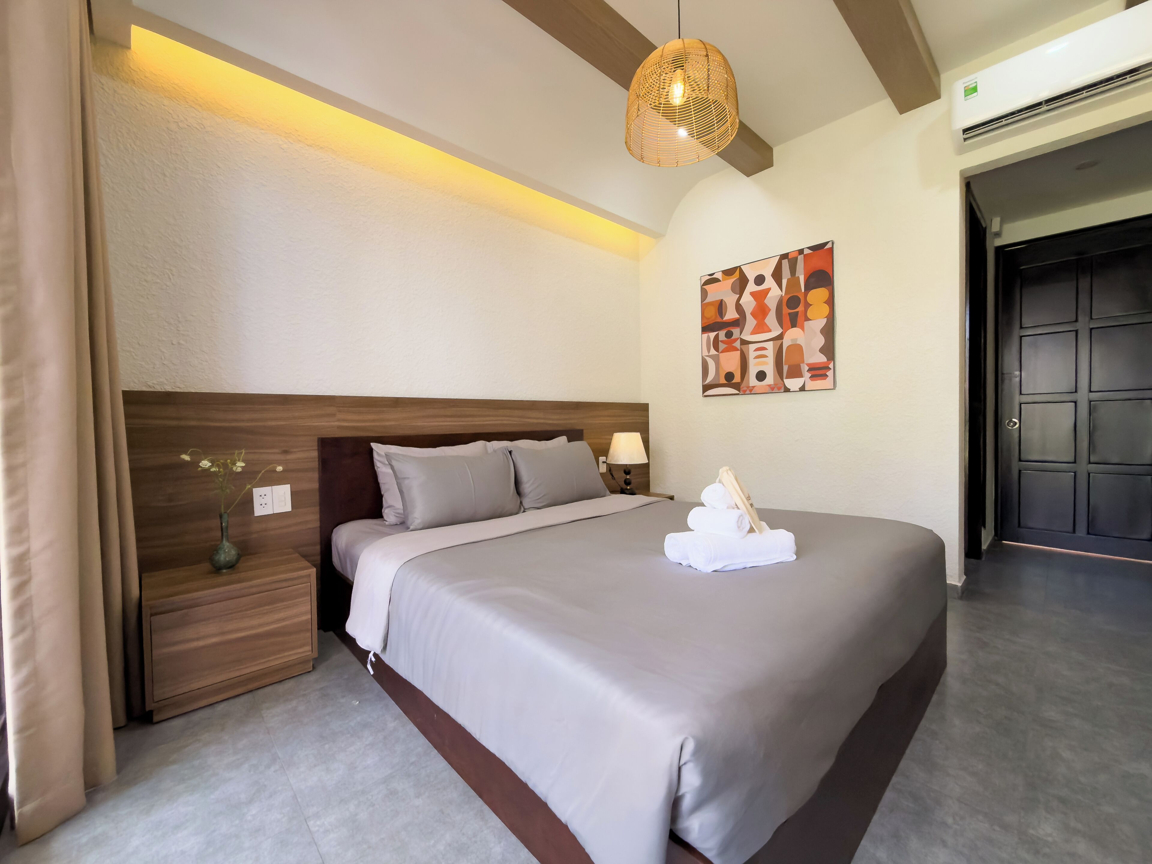 A neatly made bed with a headboard, a bedside table with a lamp, a wall-mounted abstract painting, and a door in the background.
