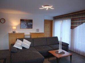 Ein Wohnzimmer mit einer Eckcouch, einem kleinen Tisch mit Kerzen, einem Gemälde an der Wand und einem Fenster mit Vorhängen.