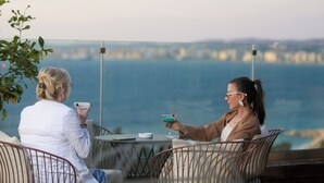 Två personer sitter vid ett bord med utsikt över havet. På bordet står ett glas med dryck och en cigarett.
