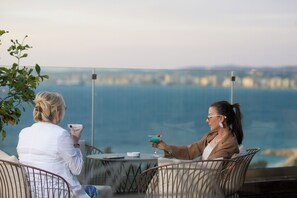 Zwei Personen sitzen an einem Tisch mit Blick auf das Meer, ein Getränk und eine Zigarette stehen auf dem Tisch.