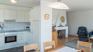 A modern kitchen with white cabinets, a microwave, and an oven. There is a dining area with a table and chairs, and a fireplace with a mantle and decorations. - Capol-Garten, (Flims Dorf), 2567 by Interhome (Flims)