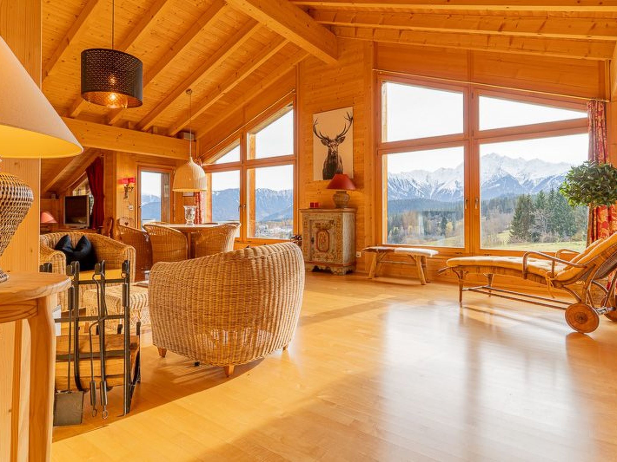 A wooden interior with a high ceiling, large windows, a wicker sofa, a dining table, and a view of mountains.