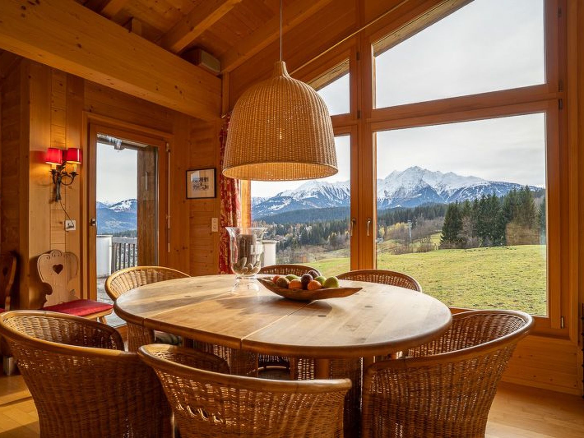 Une salle à manger en bois, avec une grande table et des chaises en osier, un luminaire suspendu et une vue sur les montagnes par de grandes fenêtres.