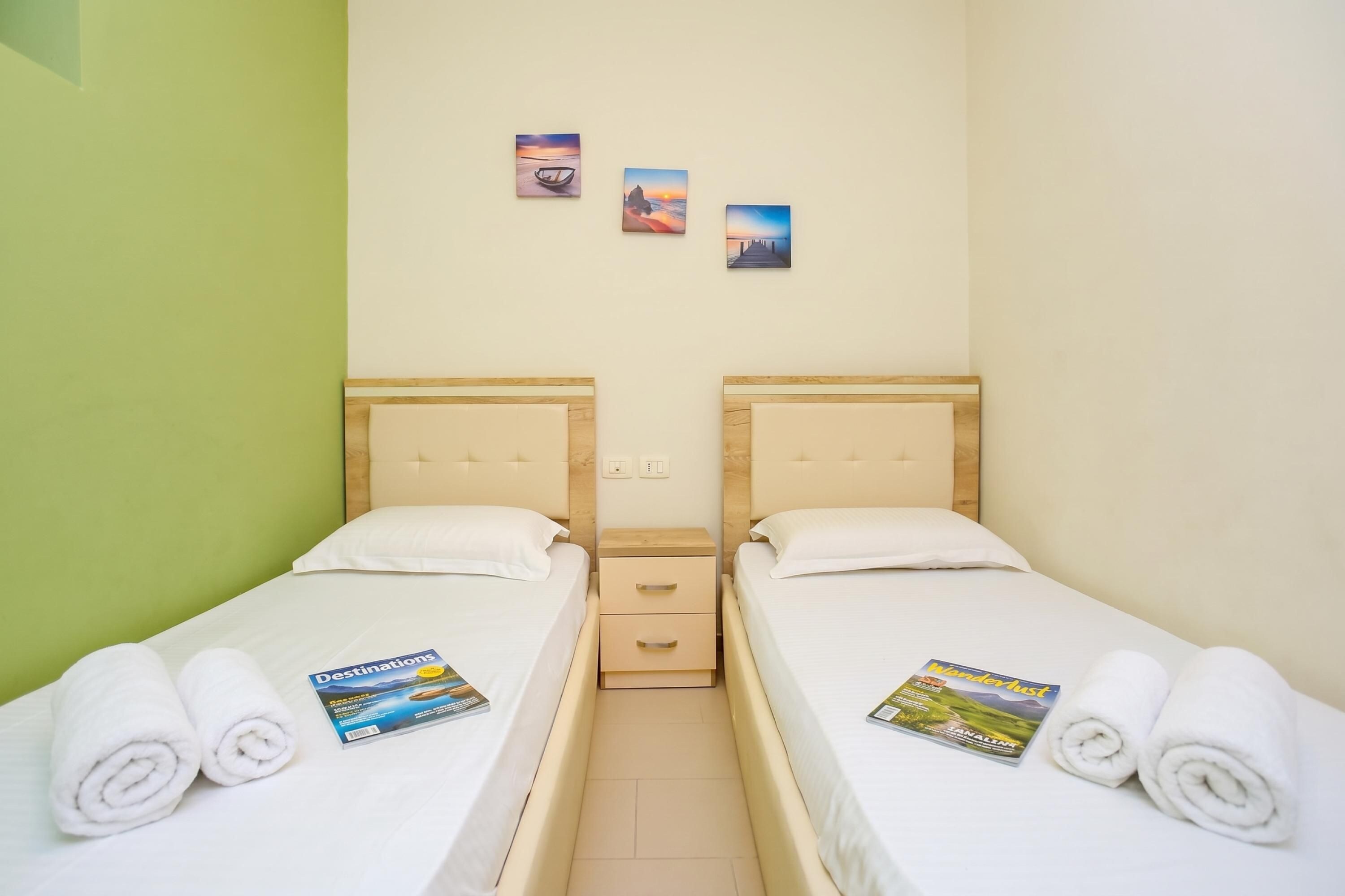 Habitación doble con dos camas, cada una con una almohada y una toalla enrollada. Hay dos cuadros colgados en la pared y una mesita de noche con cajón entre las camas.