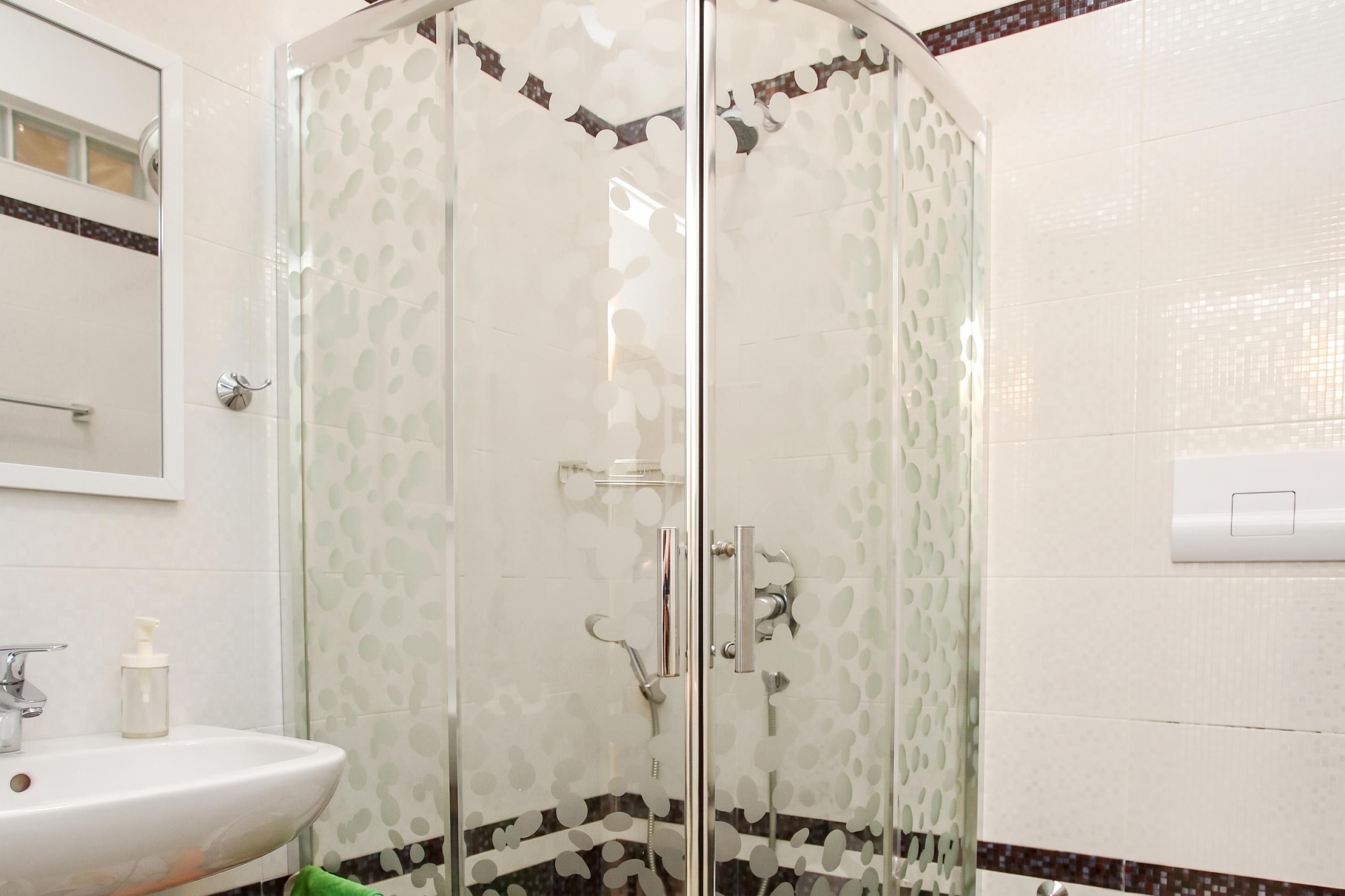 Een douche met glazen wanden, een witte betegelde muur en een spiegel in de hoek.