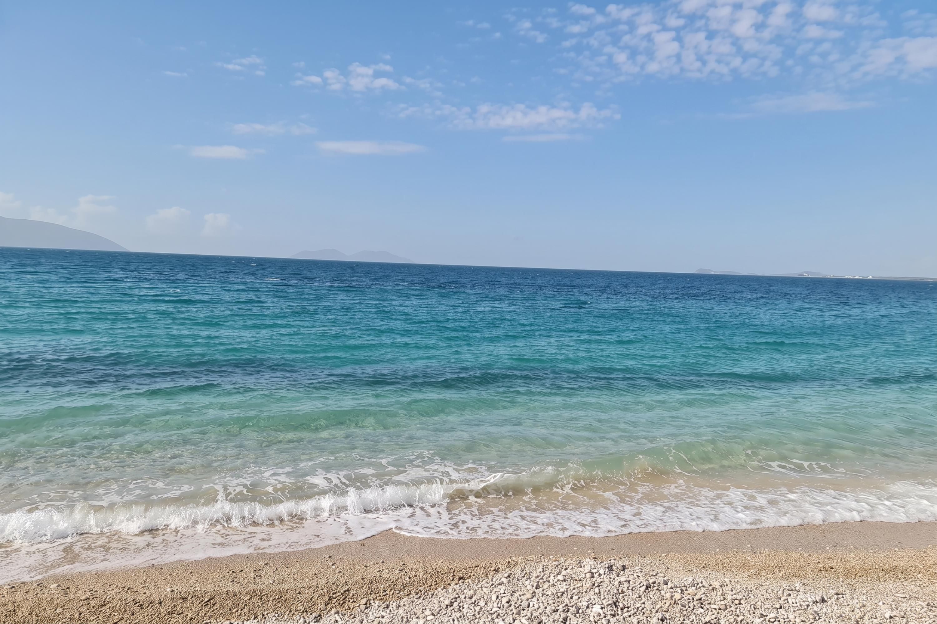 Une plage à l’eau claire, avec un rivage de galets et des montagnes qui se dessinent à l’horizon.