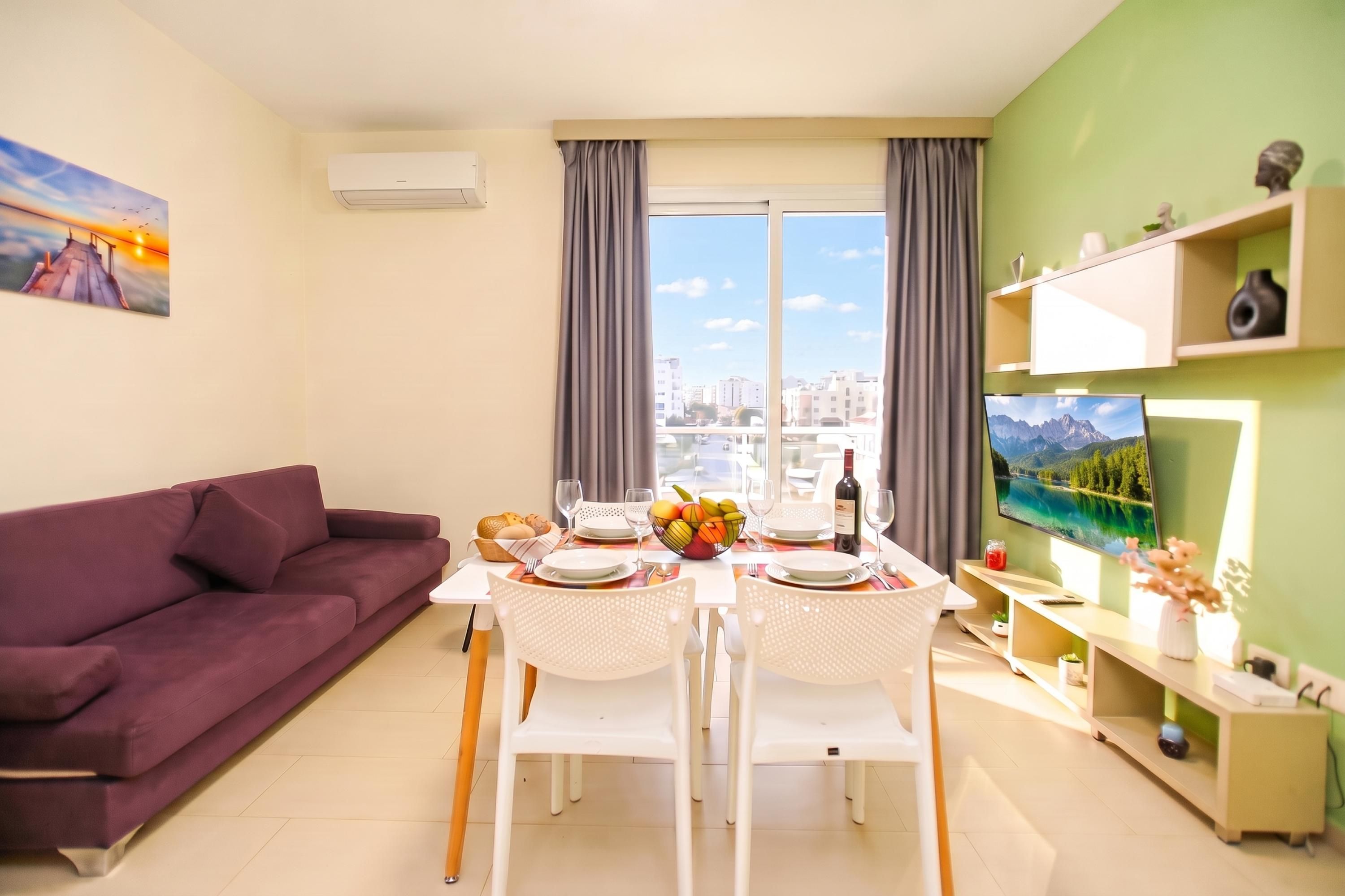 A modern living room with a sofa, dining table set for two, and a television on the wall.