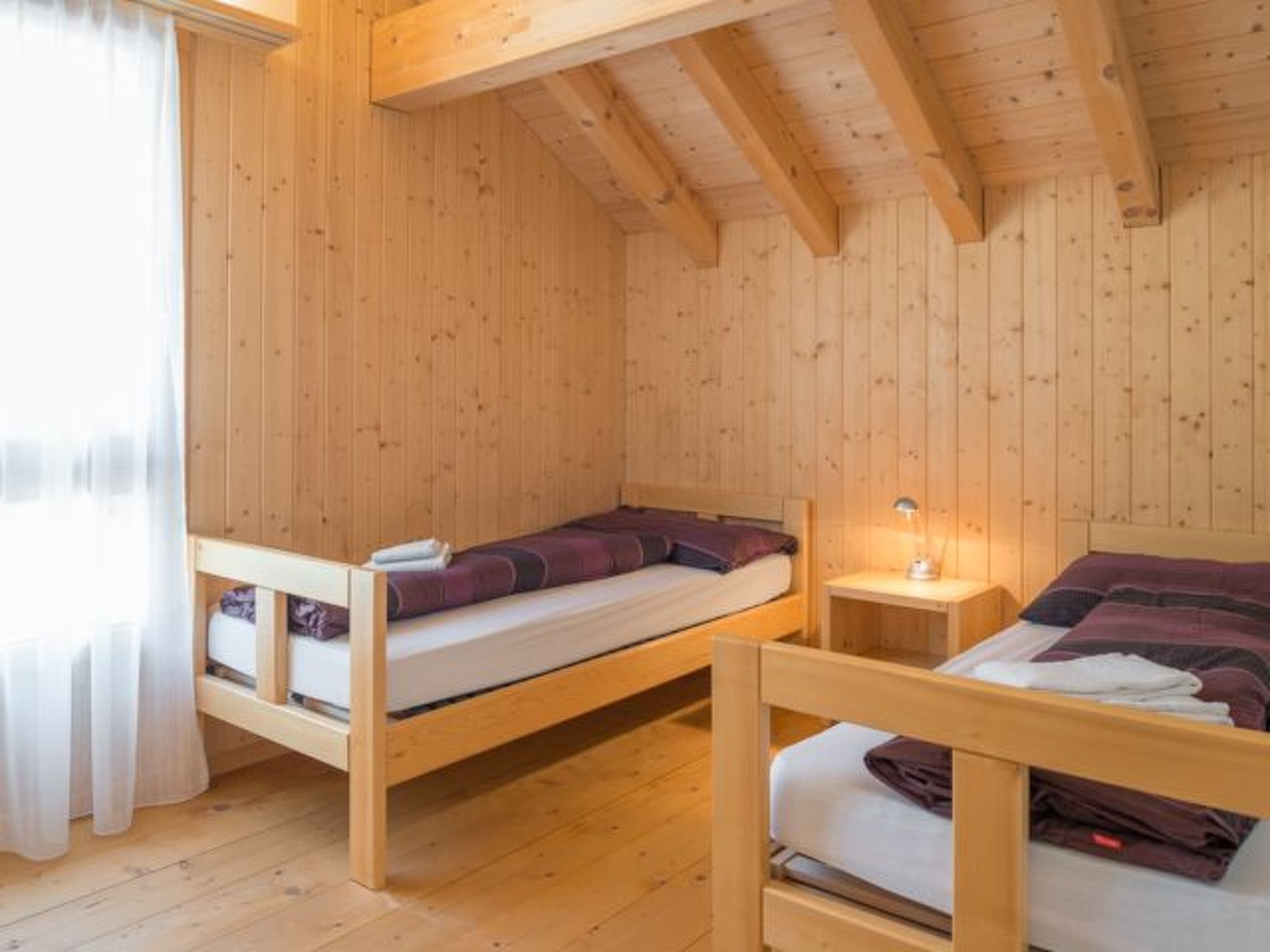 A wooden cabin with two bunk beds, a small bedside table with a lamp, and a window with curtains.