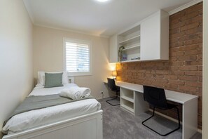 Un dormitorio con cama, escritorio y silla. Hay una ventana con persianas, una lámpara y una pared de ladrillo.