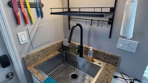 Une cuisine dotée d’un évier en acier inoxydable, d’un plan de travail en granit et d’un robinet noir. On y trouve des couteaux colorés rangés sur un présentoir, une serviette accrochée à la porte et un panneau encadré au mur.