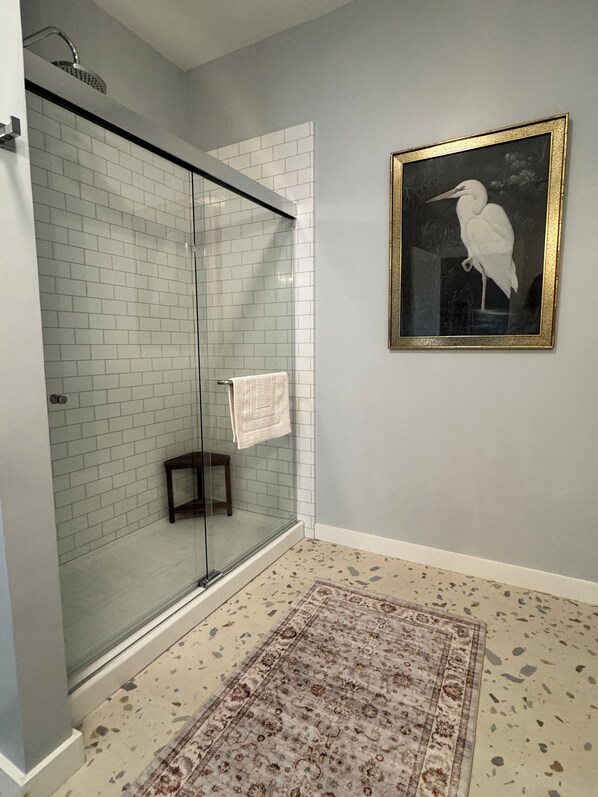 A bathroom with a glass shower enclosure, a patterned rug, a framed picture on the wall, and a small wooden stool. - Lovely 1-bedroom MCM apartment with AC near downtown New Bern (New Bern)