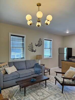 A living room with a sofa, coffee table, and armchairs, a chandelier, and a kitchen area with a refrigerator.
