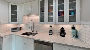 Una cocina moderna con armarios blancos, lavavajillas de acero inoxidable, tostadora empotrada, cafetera y licuadora.