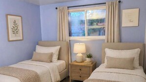 A bedroom with two beds, a nightstand with a lamp, a framed picture, and a window with curtains. - Step into The Guest House… stay awhile, where comfort meets elegance. (Inkster)