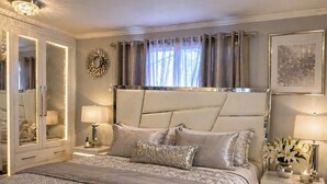 A bedroom with a large bed, a wardrobe, a nightstand with a lamp, and a decorative mirror. - Step into The Guest House… stay awhile, where comfort meets elegance. (Inkster)