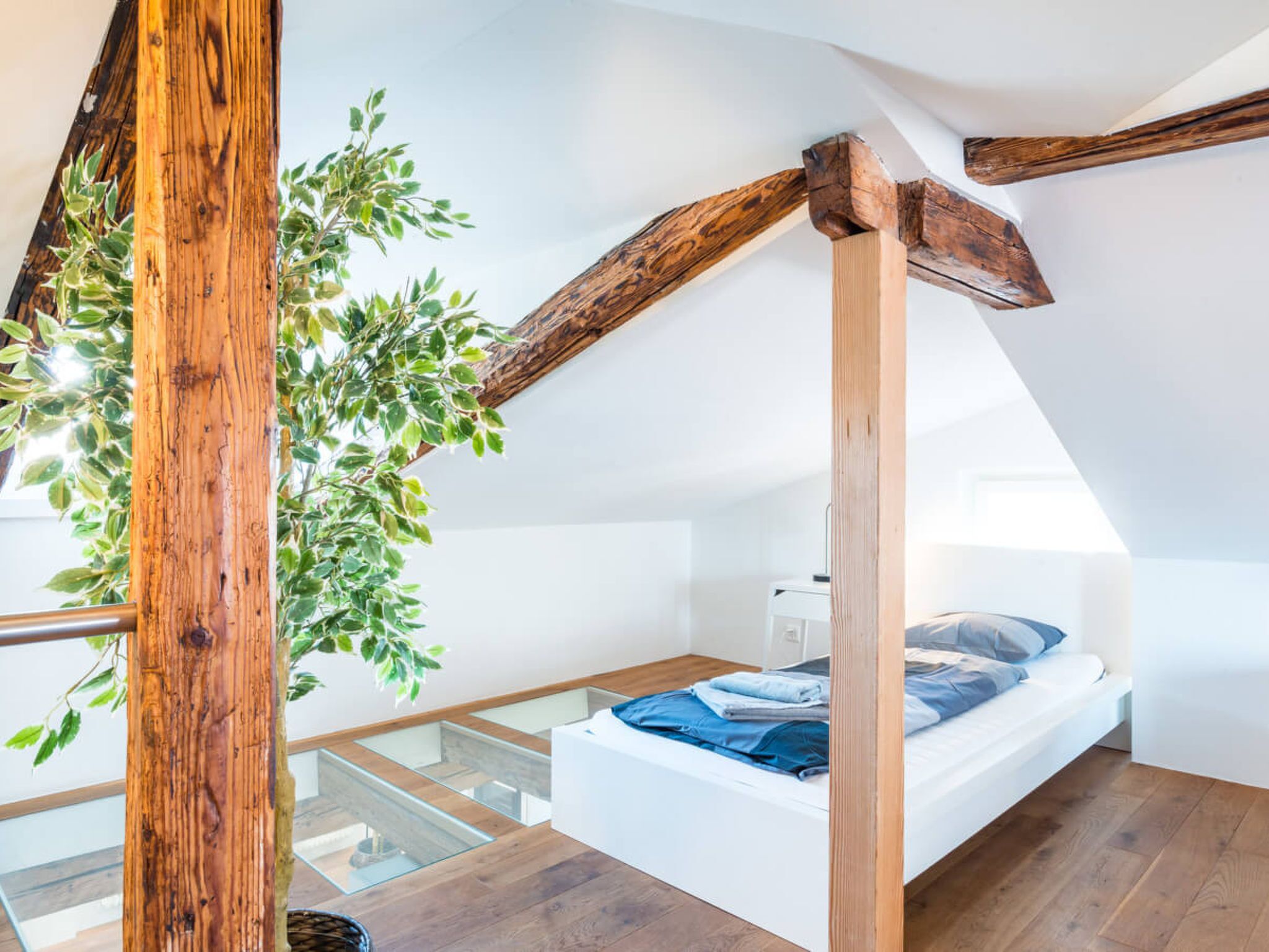 Une chambre avec un plafond en pente, des poutres en bois, un lit et une rambarde en verre.