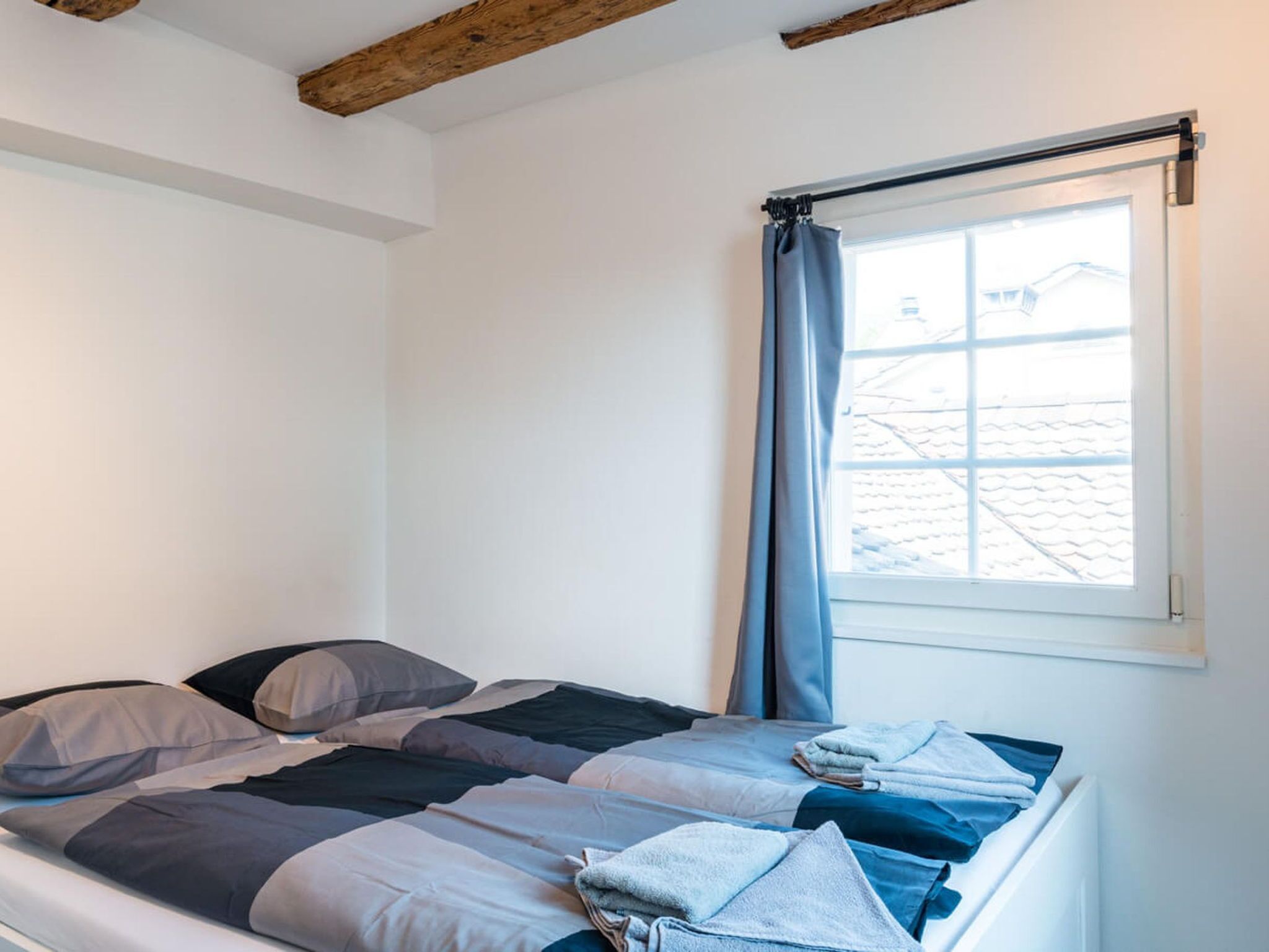 Un lit simple avec une couverture à rayures et des oreillers, une fenêtre avec un rideau et un plafond avec une poutre en bois.