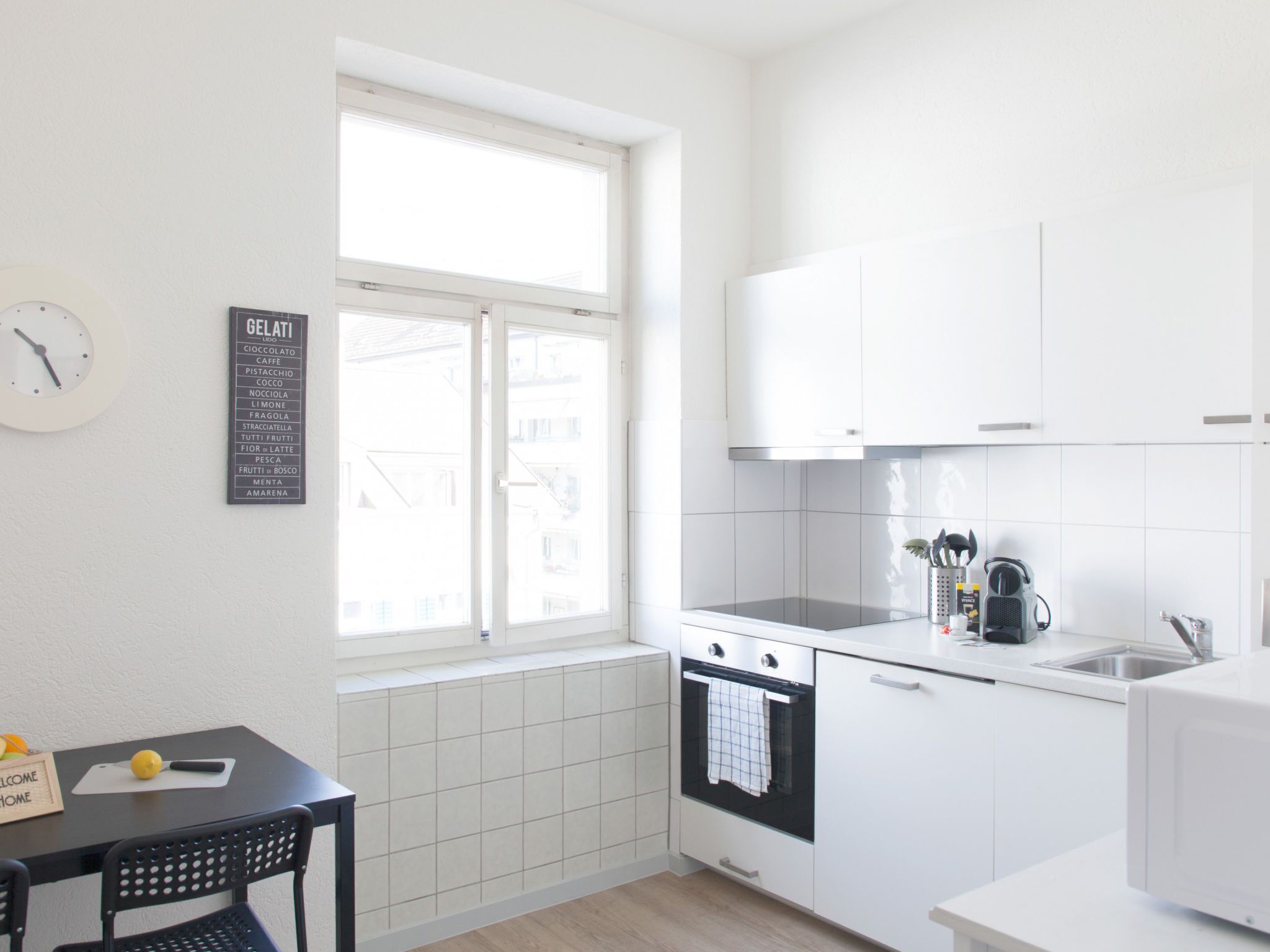 Une cuisine moderne avec des meubles blancs, une cuisinière noire et une fenêtre donnant sur les bâtiments environnants.