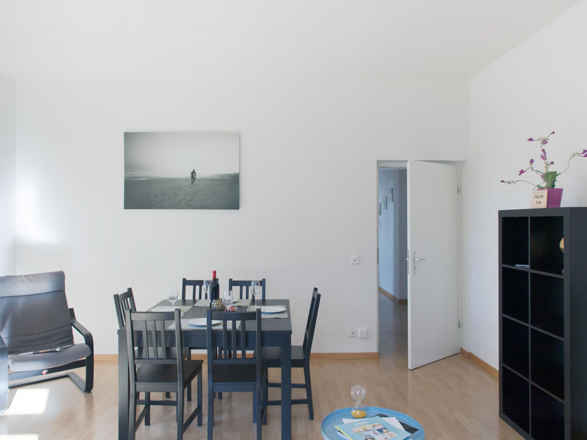Un coin repas avec une table dressée pour deux personnes, une chaise noire et une petite table d’appoint sur laquelle se trouvent un sablier et un plateau.