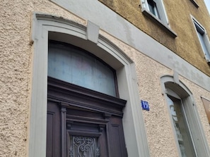 Eine Gebäudefassade mit einer dunkelholzigen Tür, einem Fenster mit dekorativem Rahmen und einem blauen Schild mit der Nummer 19.