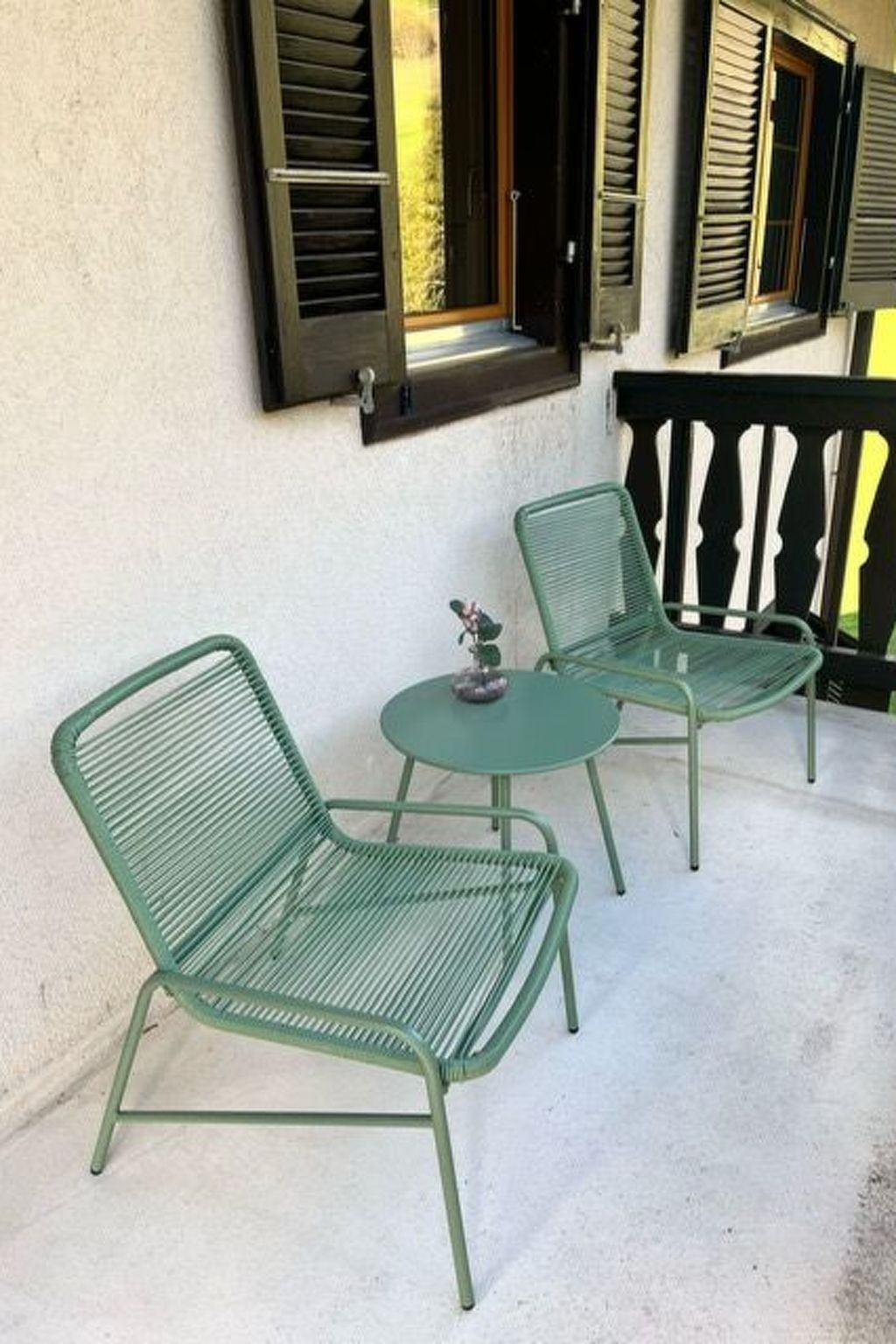 Zwei grüne Metallstühle und ein kleiner runder Tisch auf einem Balkon mit weißer Wand und schwarzen Fensterläden.