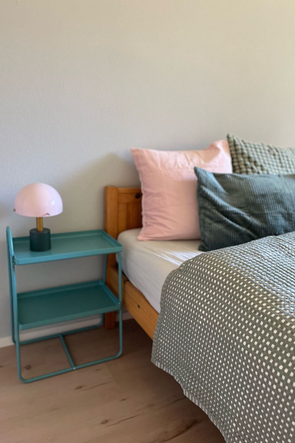 A bed with a green patterned bedspread, a teal nightstand with a lamp, and a wooden headboard.