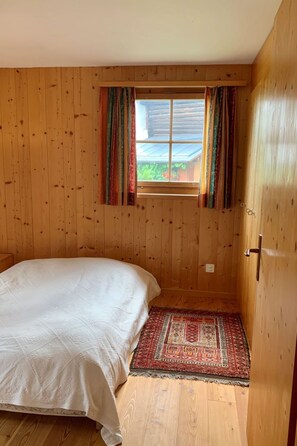 Een kamer met houten wanden, een bed met een witte dekbedovertrek, een raam met gordijnen en een kamerbreed tapijt met een patroon.