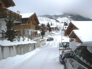Eine verschneite Straße mit Holzhäusern, einem Auto und einem Berg im Hintergrund.