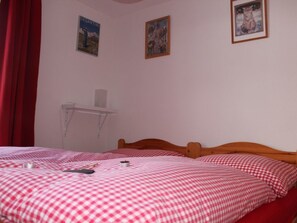 Ein Zimmer mit zwei Betten, jeweils mit einer rot-weiß karierten Tagesdecke, einem kleinen Tisch und zwei gerahmten Bildern an der Wand.