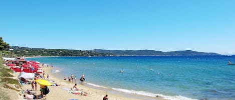 A beach with people sunbathing and swimming, red umbrellas, and a clear blue sky.