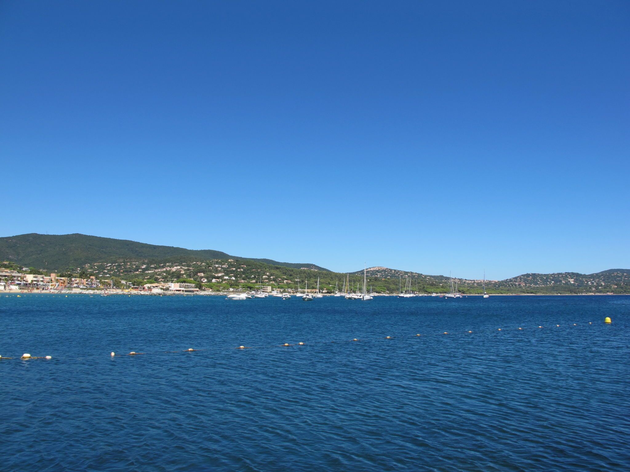 Blick auf die Küste mit Booten, klarem, blauem Wasser und einer hügeligen Landschaft im Hintergrund.