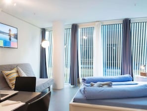 Ein Hotelzimmer mit Bett, Schreibtisch mit Laptop, Stuhl, Sofa und einem großen Fenster mit Vorhängen.