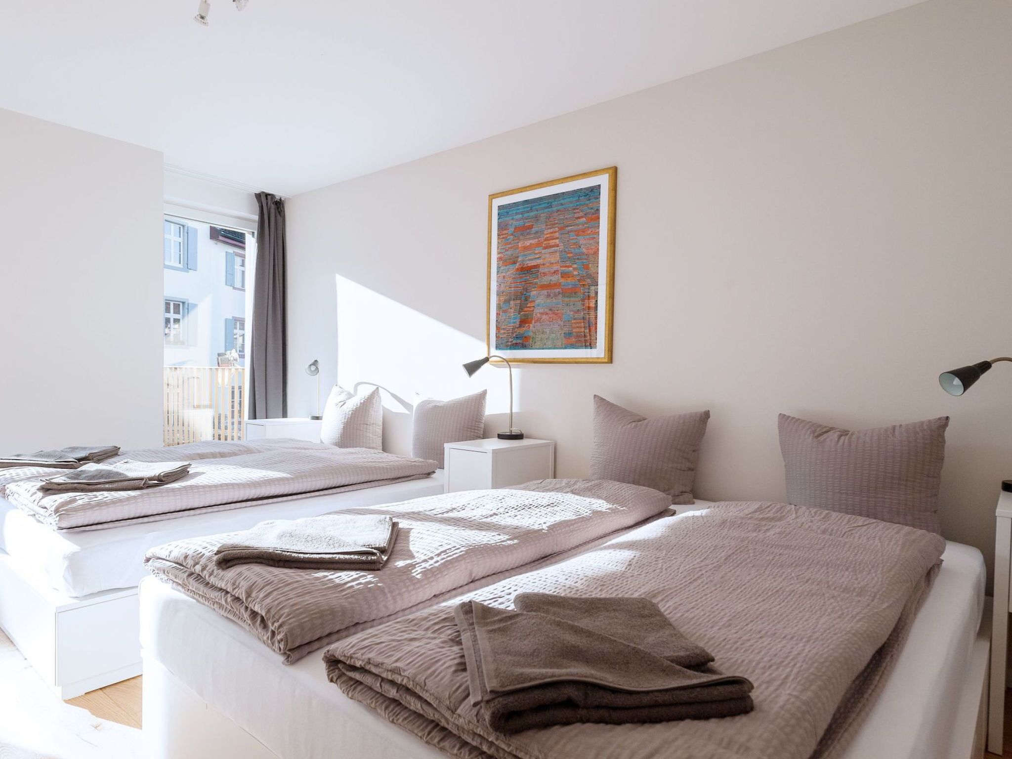 A hotel room with two beds, a large window, a painting, and a bedside lamp.