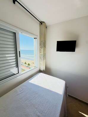 Un dormitorio con cama, ventana con persianas, cortina, televisor de pantalla plana en la pared y vista a la playa.