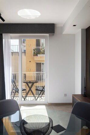 A modern room with a glass table, chairs, a sofa, a sliding glass door to an outdoor balcony, and a view of a building with balconies.