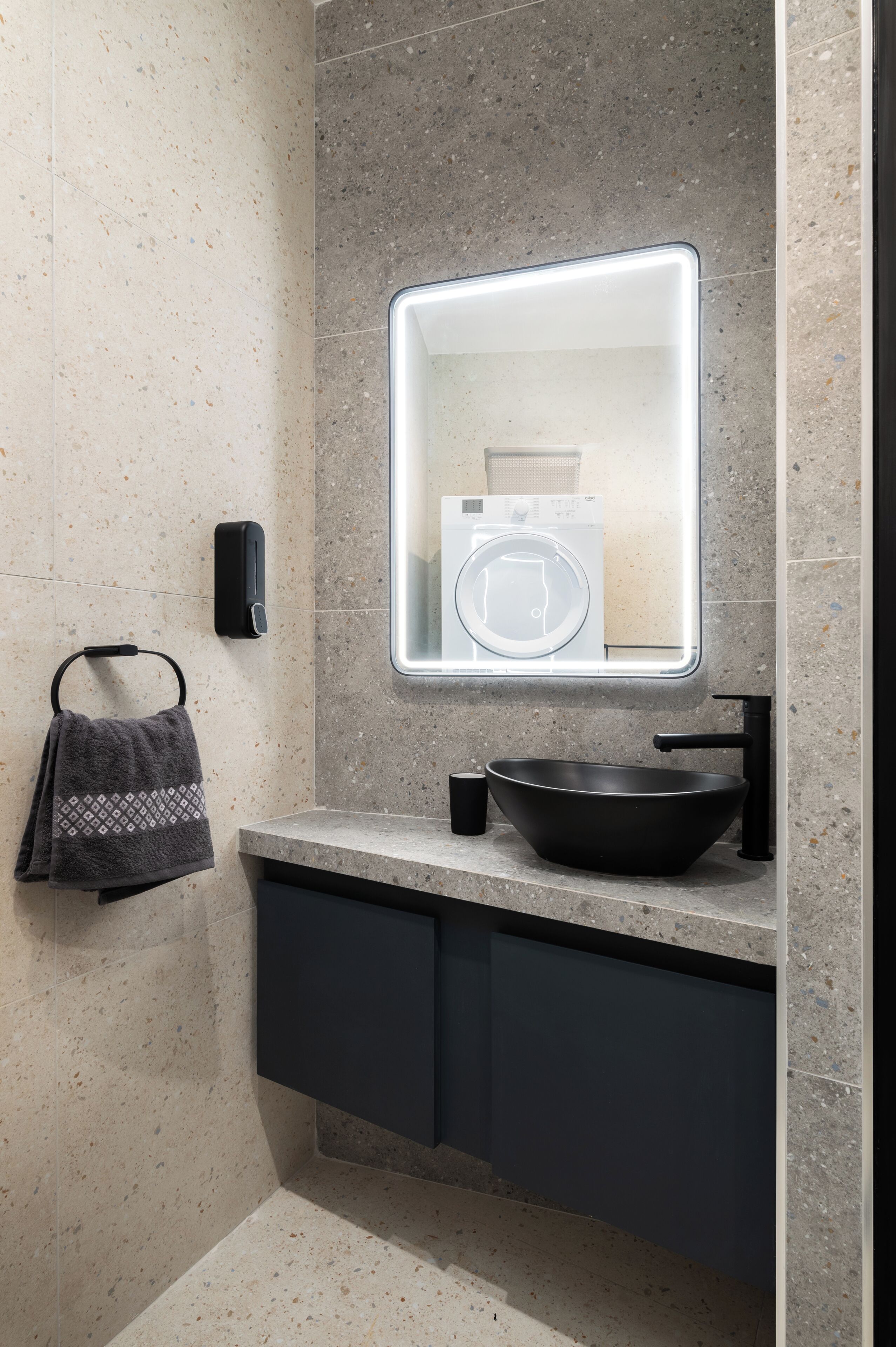Een moderne badkamer met een vrijstaand wastafelmeubel, een handdoekrek, een zeepdispenser en een spiegel met ingebouwde verlichting.