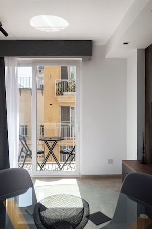 A modern room with a glass table, chairs, a sofa, a sliding glass door to an outdoor balcony, and a view of a building with balconies. - Modern 1BR APTS near Manoel & Tigne (Gżira)