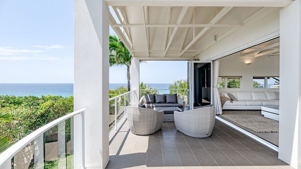 Varanda com vista para o oceano, um sofá branco, cadeiras de vime e uma porta de vidro que dá para uma sala de estar.