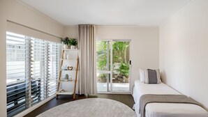 Quarto com cama, janela com persianas, planta e vista para área verde lá fora.