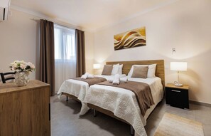 Ein Hotelzimmer mit zwei Betten, einem Schreibtisch, einem Sessel, einer Vase mit Blumen, einem Bild an der Wand und einem Nachttisch mit einer Lampe.