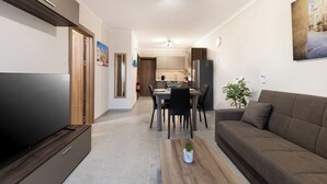 A modern living room with a flat-screen TV, a sofa, a dining table with chairs, and a kitchen in the background. - Luxury 1BR Stay at The Podium - Marina (Msida)