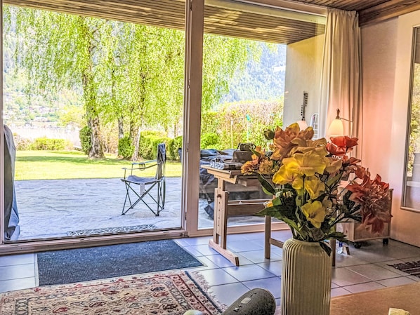A room with a large sliding glass door leading to a garden, a dining table with chairs, and a vase with flowers.