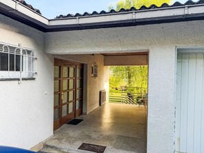 Une entrée abritée, dotée d’une porte en bois, d’un portail en métal et d’un sol en béton, qui mène à une zone herbeuse.