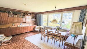 Una camera accogliente con pavimento in legno, un tavolo da pranzo con sedie, una libreria e una finestra vista sul verde.