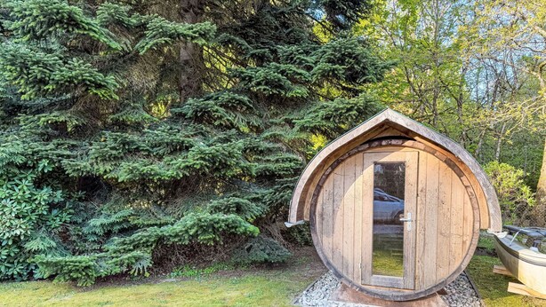 Una struttura a forma di botte in legno, con una porta, circondata da erba e alberi.