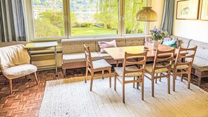 Un'area pranzo con tavolo e sedie in legno, una panca e vista su un giardino attraverso ampie finestre.