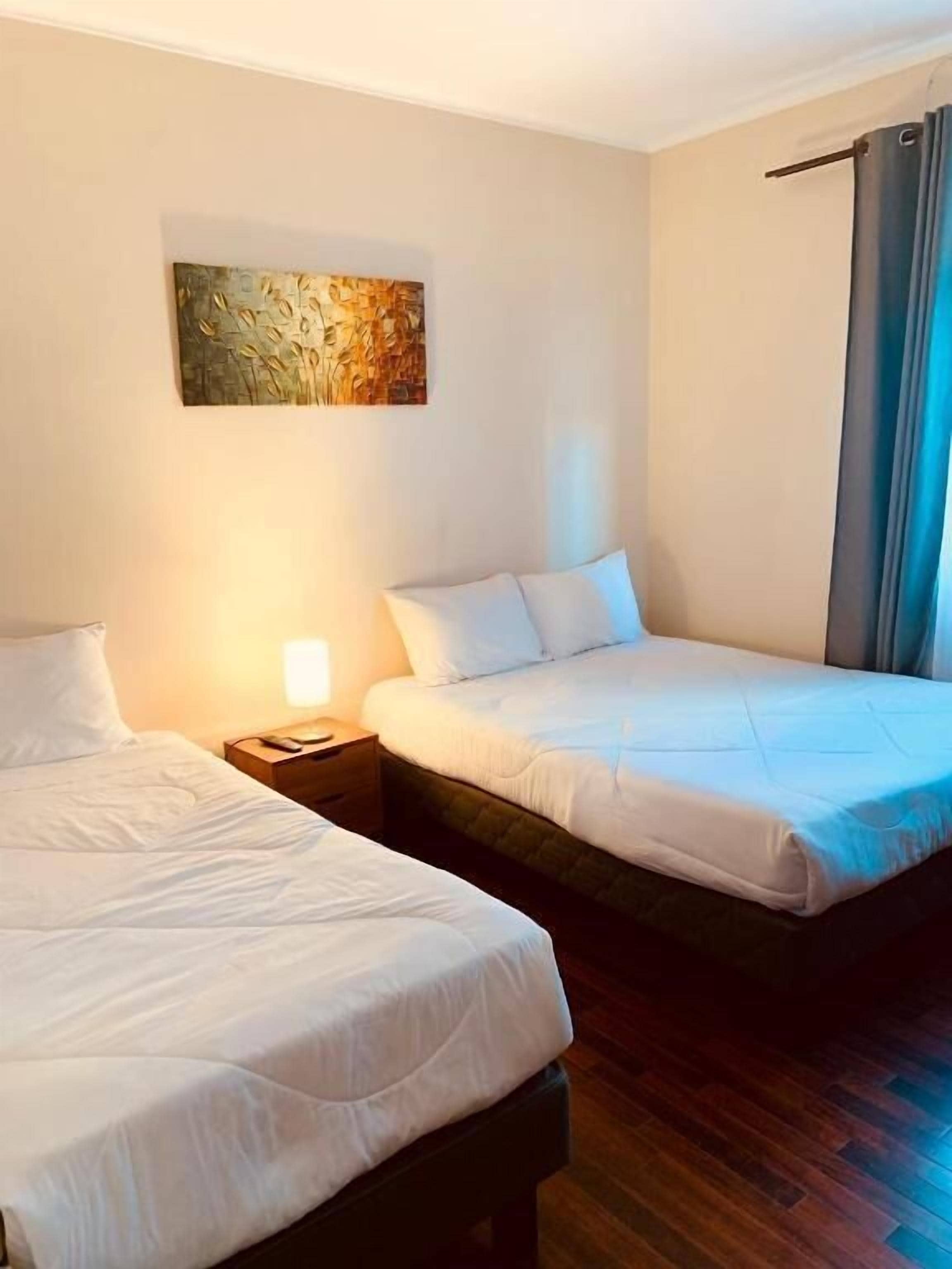 A hotel room with two beds, a nightstand, a lamp, and a painting on the wall.