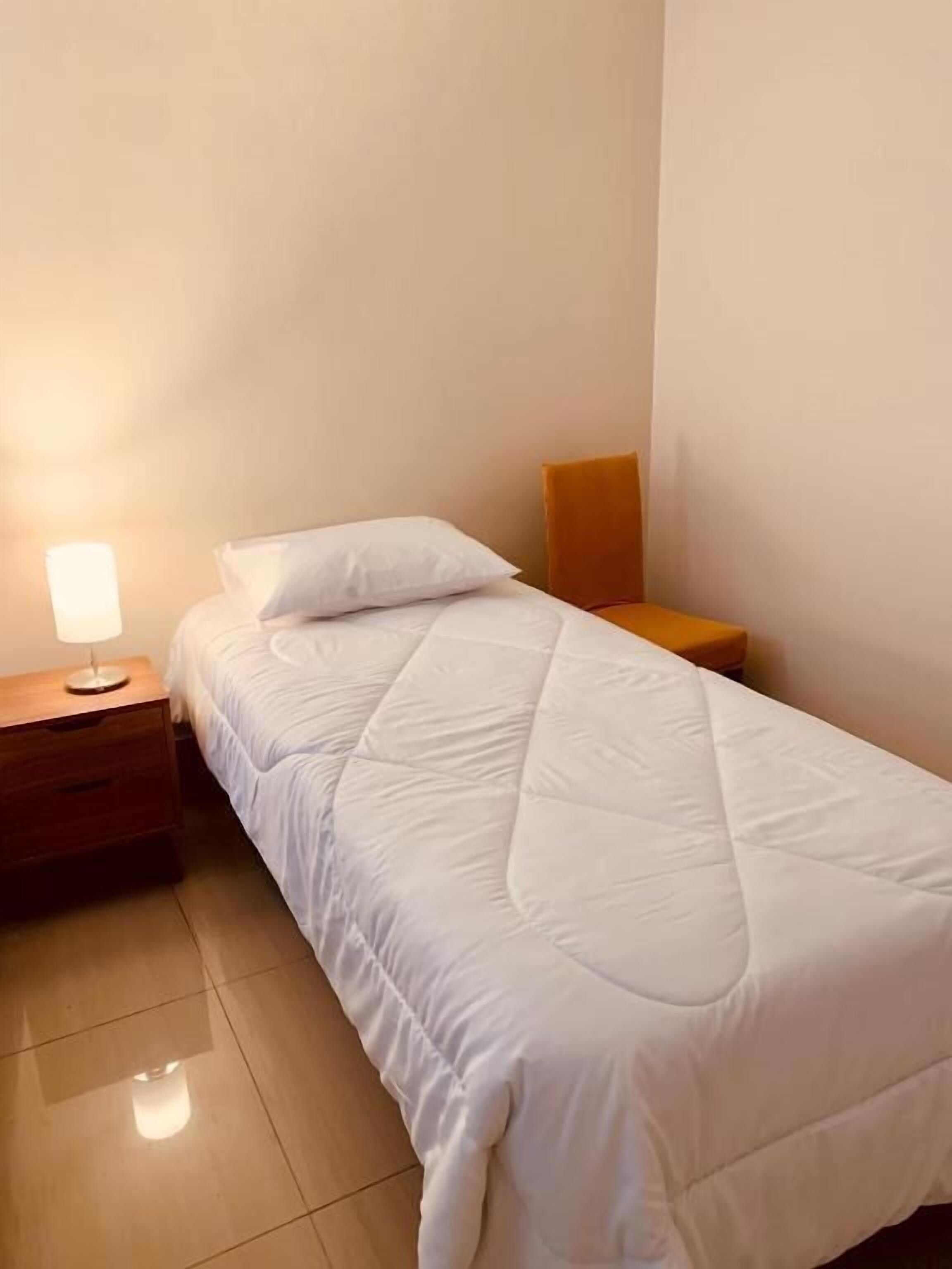 Un lit simple avec une couette blanche, une table de chevet en bois avec une lampe et un mur uni.
