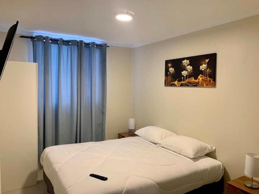 A hotel room with a bed, curtains, a painting, a nightstand, and a lamp.